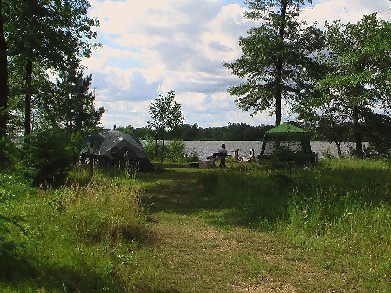 A tent and a shade canopy on a grassy walk-in campsite among trees beside a lake in a public state park.